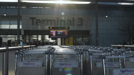 Reihe-Von-Gepäckwagen-Vor-Dem-Heathrow-Terminal-3-Gebäude-Mit-Abflugtafel-Der-Fluggesellschaft-Mit-Sonneneruptionen