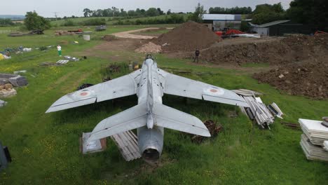 Aerial-view-descending-behind-neglected-Hawker-hunter-fighter-jet-among-discarded-junk-in-field