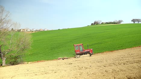 Small-tiny-red-tractor-driving-uphill-pull-triple-rolling-system-equipment-preparing-the-land-in-agricultural-natural-farm-during-a-sunny-day-of-spring