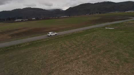 white-2019-toyota-corolla-hatchback-sports-car-sedan-driving-along-country-farm-road-surrounded-by-grass-brown-fields-mountains-in-farmland-Abbotsford-BC-Aerial-tracking-follow