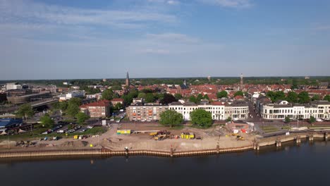 Construction-site-of-medieval-city-seen-from-above-with-inland-shipping-large-cargo-vessel-leaving-ripple-waves-on-river-IJssel-passing-IJsselkade-boulevard-cityscape-of-tower-town-Zutphen