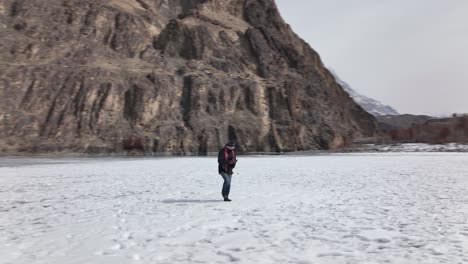 Männchen-Zu-Fuß-über-Den-Zugefrorenen-Khalti-See,-Warm-Eingepackt-Mit-Mütze-Und-Schal-Und-Schneebedeckten-Bergen-Im-Hintergrund