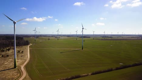 Aerial-view-of-wind-turbines-generating-renewable-energy-in-the-wind-farm,-sunny-spring-day,-low-flyover-over-green-agricultural-cereal-fields,-countryside-roads,-wide-angle-drone-shot-moving-forward