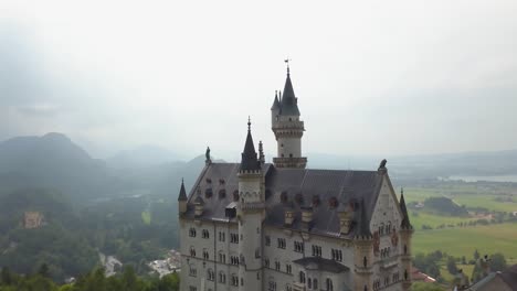 Aerial-orbit-of-fairytale-Neuschwanstein-Castle-on-a-hill-surrounded-by-woods,-Pollat-river-and-village-in-background-on-an-overcast-day,-Bavaria,-Germany