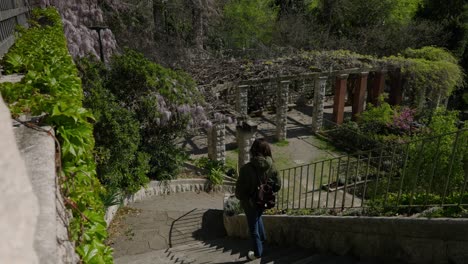 Trieste,-Italy,-03-11-2022,-Young-woman-walking-down-a-staircase-in-a-historic-garden-with-wisteria-flowers-and-pergolas