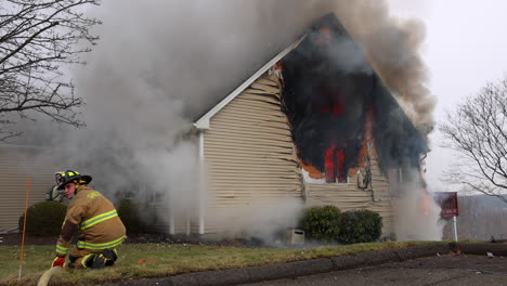 A-Firefighter-Kneels-Down-Holding-onto-a-Fire-Hose-Outside-of-a-Burning-Residential-House