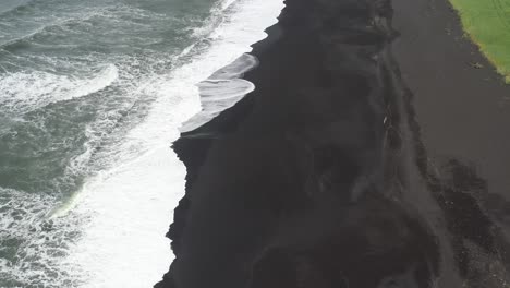 Black-sand-beach-with-waves-in-Vik,-Iceland-with-drone-video-above-moving-back