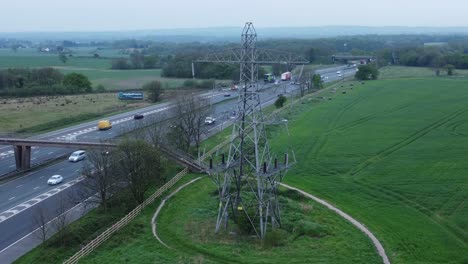Vehicles-on-M62-motorway-passing-pylon-tower-on-countryside-farmland-fields-aerial-view-slowly-rising-shot