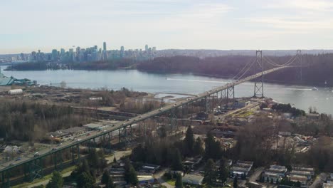 Fliegen-über-Die-Lions-Gate-Bridge-Und-Blick-Auf-Eine-Straßenwut-Von-Vorbeifahrenden-Fahrzeugen,-Das-Ruhige-Flussufer-Und-Die-Strukturelle-Metropole-In-Vancouver,-Kanada