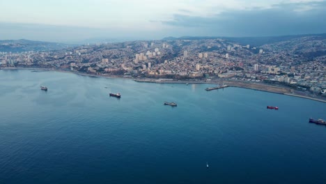 Aerial-dolly-in-of-container-cargo-ships-sailing-near-the-coastline-and-Sea-Port,-Valparaiso-hillside-city,-Chile