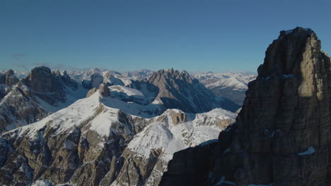 Aerial-pull-back-view-from-snowy-mountain-alps-terrain-to-reveal-sunlit-Tre-Cime-stunning-peaks