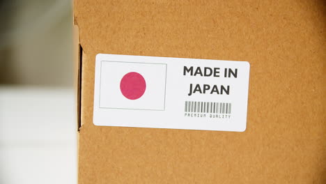 Manos-Aplicando-La-Etiqueta-De-La-Bandera-Hecha-En-Japón-En-Una-Caja-De-Cartón-De-Envío-Con-Productos