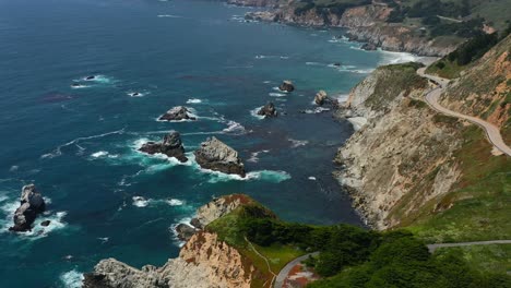 Wide-aerial-view-of-the-winding-curvy-highway-road-along-Route-1-on-the-coast-of-Big-Sur-California-with-waves-crashing-into-the-large-rocks-in-the-Pacific-Ocean