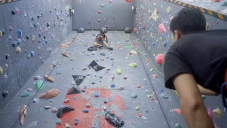Rock-climber-on-indoor-wall-belayed-by-friend-at-bottom