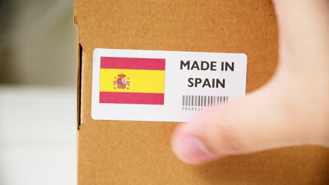 Manos-Aplicando-La-Etiqueta-De-La-Bandera-Hecha-En-España-En-Una-Caja-De-Cartón-De-Envío-Con-Productos