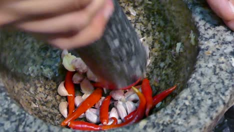 Asian-Hand-Using-Stone-Mortar-and-Pestle-Crushing-Garlic-and-Red-Chili-Pepper,-Close-Up,-Slow-Motion
