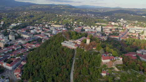Aerial-view-flying-high-around-historic-Glockenturm-tower-overlooking-Graz-Schloßberg-dense-hilltop-woodland-cityscape