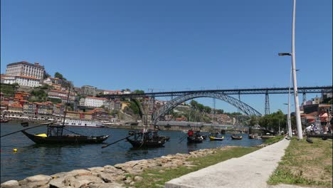 Zeitraffer-Des-Flusses-Duoro-Und-Der-Brücke-Ponte-Luis-Während-Eines-Atemberaubend-Klaren,-Heißen-Europäischen-Sommertages-Mit-Tourbooten,-Die-Im-Forgronud-Schwimmen,-Und-Touristen,-Die-Entlang-Der-Wasserkante-Spazieren,-Porto,-Portugal