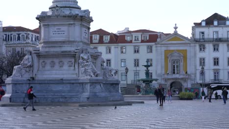 People-walking-at-Praca-do-Russio-near-statue-of-King-Dom-Pedro-IV,-public-square-beautiful-Portuguese-pavement-in-Lisbon