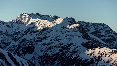 Low-angle-shot-of-beautiful-snow-covered-white-winter-mountain-peak-in-evening-time-with-sun-going-down-along-the-side-in-timelapse
