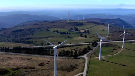 Windkraftanlagen-Drehen-Sich-In-Den-Bergen-Mit-Weidendem-Vieh-In-Der-Nähe,-Straße-Mit-Einem-Fahrenden-Auto,-Sonniger-Tag-Und-Ein-Klarer-Blauer-Himmel