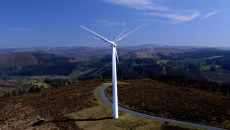 Two-wind-turbines-lined-up-spinning-their-blades-in-the-mountains-with-small-green-forests-of-trees-on-a-sunny-afternoon-of-blue-sky
