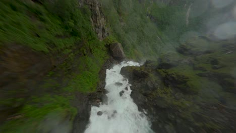 FPV-flight-down-Bjødnabykset-Waterfall-between-greened-mountains-during-rainy-day-in-Norway