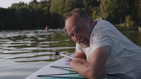 A-happy-elderly-man-leaning-on-a-paddleboard-smiles-at-the-camera