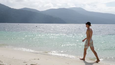 Ultrazeitlupenaufnahme-Eines-Jungen-Kaukasischen-Mannes-Mit-Kappe-Und-Badebekleidung,-Der-An-Einem-Sonnigen-Tag-In-Thailand-Am-Strand-Spazieren-Geht