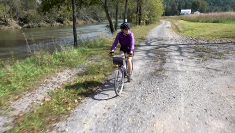 Pretty,-mature-woman-on-a-mountain-bike-riding-on-a-gravel-road-with-a-river-on-one-side-and-farmland-on-the-other