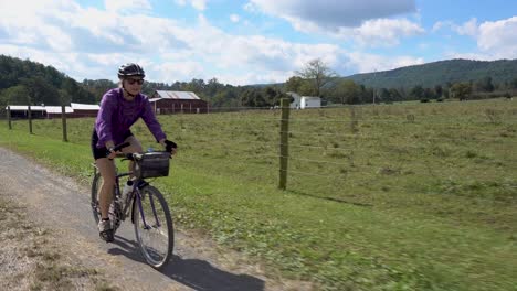 Mature-woman-with-sunglasses-biking-on-a-gravel-road-with-a-farm-in-the-distance