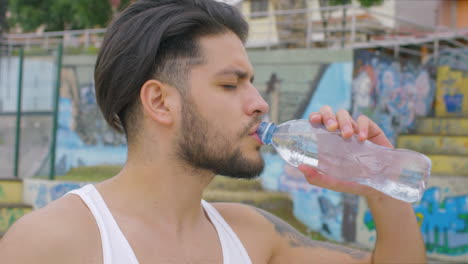 Hübscher-Kerl,-Der-In-Einem-Skatepark-Frisches-Wasser-Aus-Einer-Plastikflasche-Trinkt