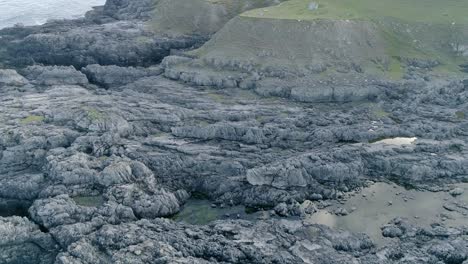 Aerial-tracking-over-a-large-swathe-of-rocky-coastline-with-crags,-inlets,-caves-and-rockpools-before-tracking-forward-over-the-grassy-hilly-coastline