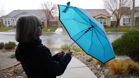 Eine-Alte-Frau,-Die-Einen-Blauen-Regenschirm-öffnet-Und-In-Einen-Wintersturm-Hinausgeht,-Während-Regentropfen-In-Zeitlupe-Fallen