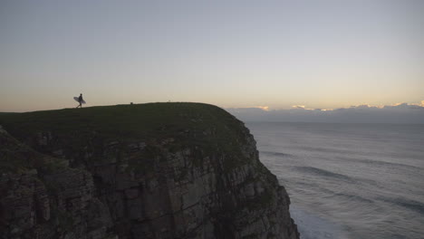 Man-hiking-out-to-edge-of-cliff-at-sunrise-with-surfboard