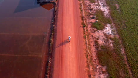 Drone-follows-biker-riding-on-dusty-dirt-road-between-farms-and-salt-plantations
