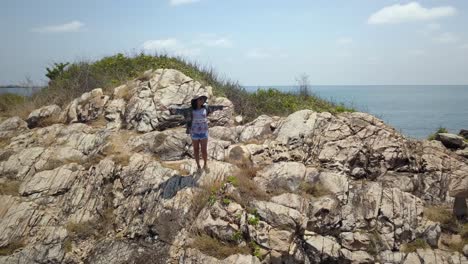 Aerial:-a-drone-flies-around-a-young-thai-woman-standing-at-a-steep-rocky-slope-,-rising-her-arms-in-the-air,-smiling-and-enjoying-her-vacations,-Thailand