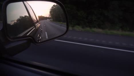 The-setting-sun,-seen-in-the-side-mirror-of-a-moving-car,-on-an-empty-road-in-a-rural-environment