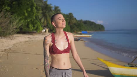 Thin-woman-with-shaved-head-standing-on-a-beach-at-the-edge-of-the-water-with-her-face-to-the-sun