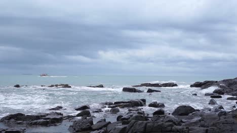 SLOWMO---Ocean-view-with-black-rocks,-waves,-boat-and-seagull-in-Bluff,-New-Zealand-on-a-cloudy-day
