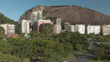 Lento-Ascenso-Aéreo-Con-Edificios-Residenciales-Y-Una-Montaña-Rocosa-Detrás-Que-Revela-Una-Carretera-A-Través-De-Un-Entorno-Verde-En-Río-De-Janeiro-Al-Atardecer