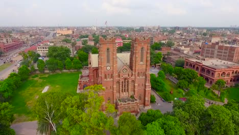 Aerial-view-of-a-beautiful-old-Church-with-city-in-the-background,-Beautiful-trees-and-grass-around-the-Church,-Pigeons-flying-over-the-Church,-People-moving-outside-the-Church
