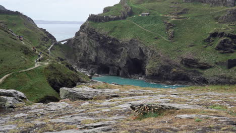 Tourists-walking-on-top-of-the-cliffs-by-the-ruins-of-Tintagel-castle-in-northern-Cornwall-with-Tintagel-beach-and-bay-below