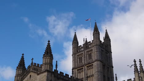 Super-Slow-Motion-Shot-of-Seagull-Flying-Through-Framer-with-Tower-of-Bath-Abbey-in-Somerset,-England-in-the-Background-on-a-Sunny-Summer’s-Day-with-Saint-George’s-Cross-Flapping-in-the-Wind