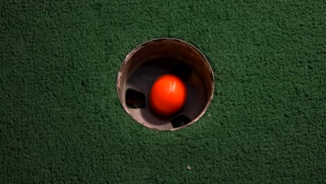 Eine-Nahaufnahme-Aus-Der-Vogelperspektive-Eines-Orangefarbenen-Minigolfballs-Fällt-In-Das-Golfloch-Und-Springt-In-Der-Tasche-Auf-Einem-Kurs