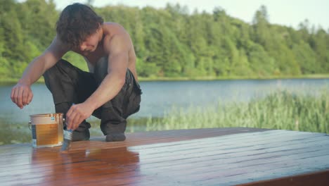 Young-carpenter-sits-in-primitive-squat-painting-roof-planking-of-wooden-boat-with-exterior-protective-paint
