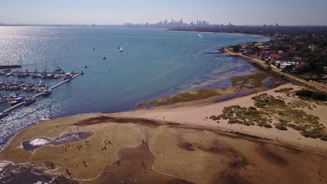 Reverse-aerial-view-over-beach-in-Port-Philip-Bay-with-Melbourne-skyline-in-background