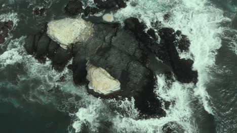 Aerial-ascending-of-rocky-formations-while-the-waves-crash-against-it-on-a-cloudy-day-in-Pichilemu,-Chile-4K