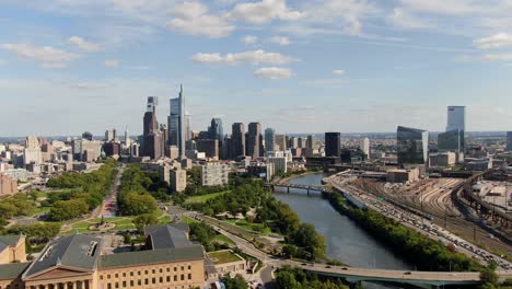Aerial-backward-flying-drone-shot-revealing-Philadelphia-Museum-of-Art-beside-Schuylkill-River-with-beautiful-Philly-skyline-against-blue-summer-sky