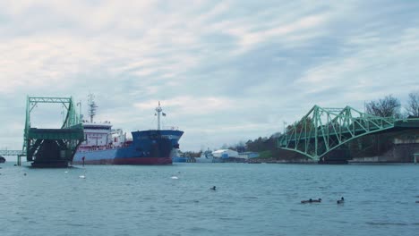Blue-cargo-ship-enters-the-port-via-an-open-Oskara-Kalpaka-swing-bridge-in-Liepaja-in-cloudy-autumn-day,-wild-ducks-in-foreground,-wide-shot